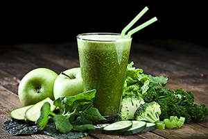 Glass of fresh green vegetable juice with two drinking straws on rustic wood table. The glass is surrounded by green vegetables like spinach, lettuce, broccoli, celery, green apples, parsley and cucumber.