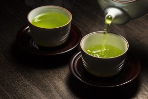 A wooden surface holding two teacups on saucers, one full of green tea, the other with green tea being poured into it from a teapot.