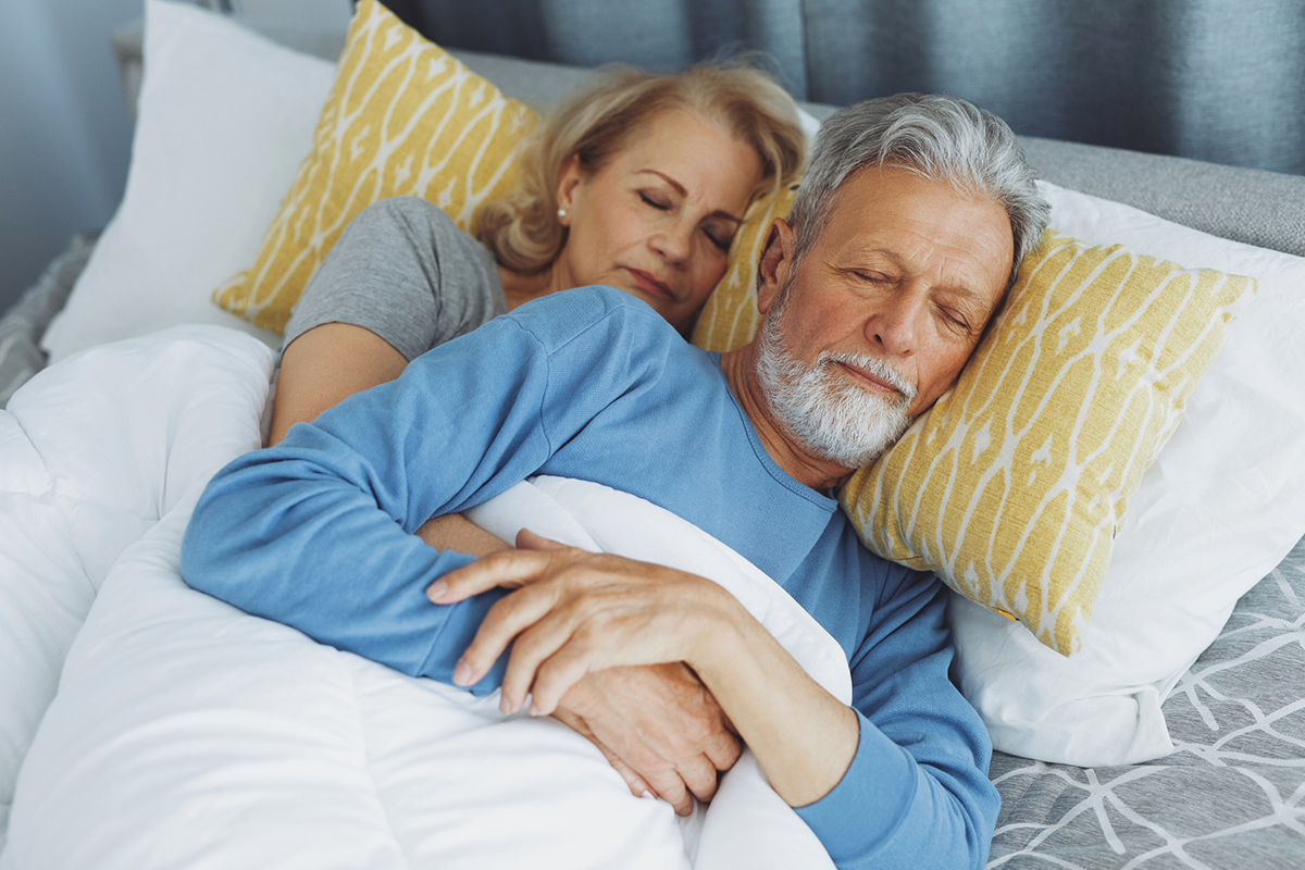 Senior couple asleep in bed - a woman with medium length blonde hair rests behind a men with short gray hair and beard. She has her arms wrapped around him and he has one hand resting atop hers. The man is in a blue long-sleeve shirt; the woman a teal short sleeve. The pillow they're resting on is yellow; the sheets are white.