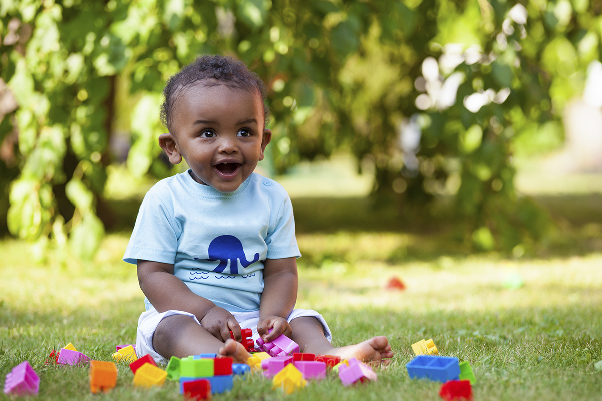 African American baby sitting up in the grass and smiling with multicolored toys strewn around him