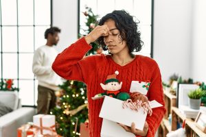 Young Hispanic woman standing by Christmas tree with decoration tired rubbing nose and eyes feeling fatigue and headache. stress and frustration concept.