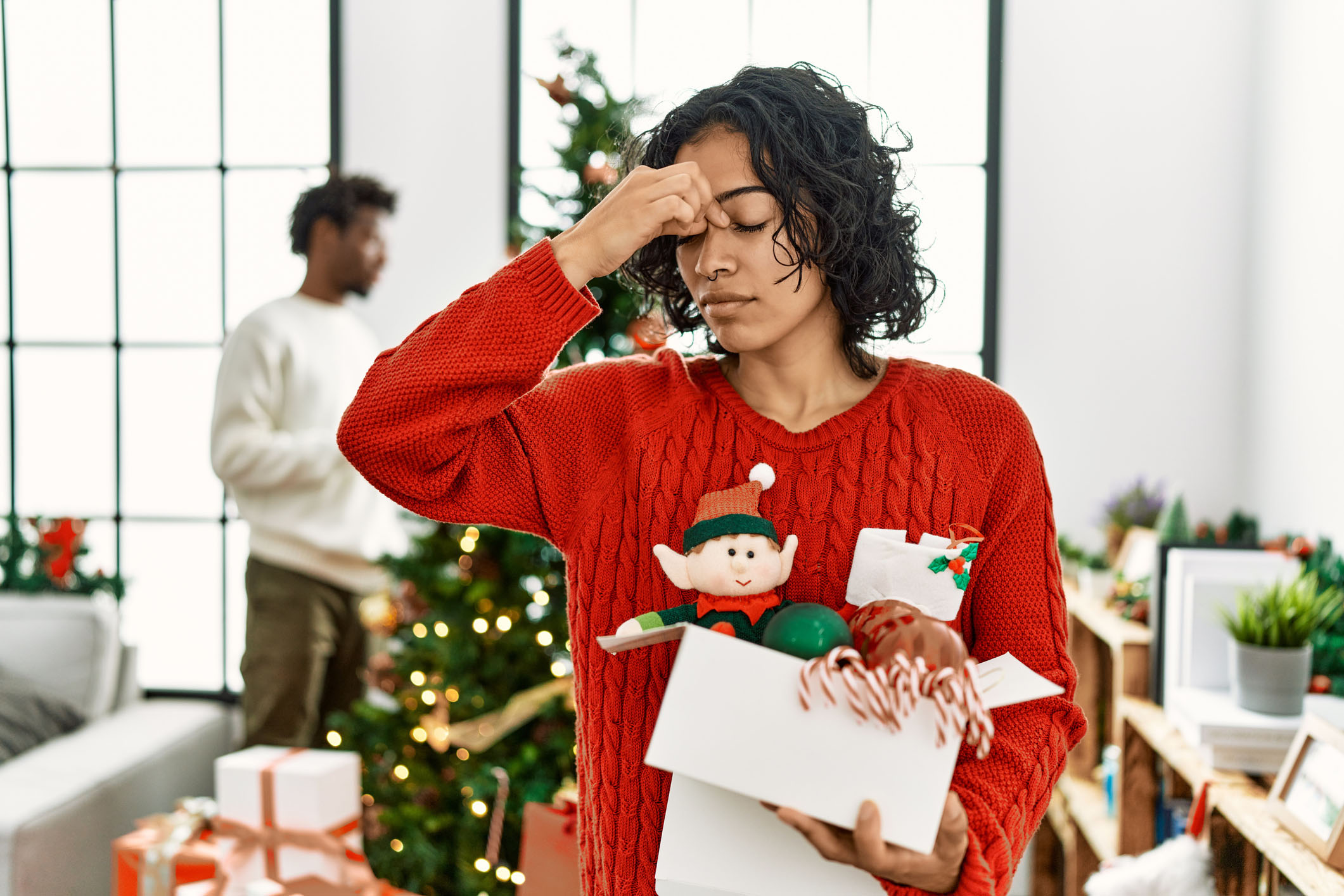 Young Hispanic woman standing by Christmas tree with decoration tired rubbing nose and eyes feeling fatigue and headache. stress and frustration concept.
