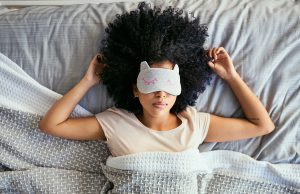 Woman with natural curly hair asleep on her back with her hands resting near her head on the pillow. She's wearing a sleep mask with cat ears and a drawing of closed eyes, and is under a woven blanket.