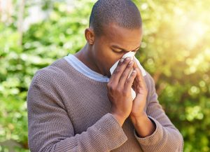 African American man in a long sleeved shirt, blowing his nose with a background of trees. Allergy concept.