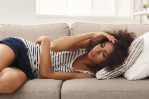 African American woman in black and white striped tank top laying on a gray couch looking ill. She has one arm over her middle and the other pressed to her head as if in pain.