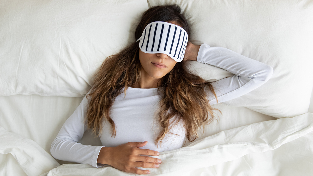 Woman with long brown hair asleep in bed with a black and white striped eye mask on her face