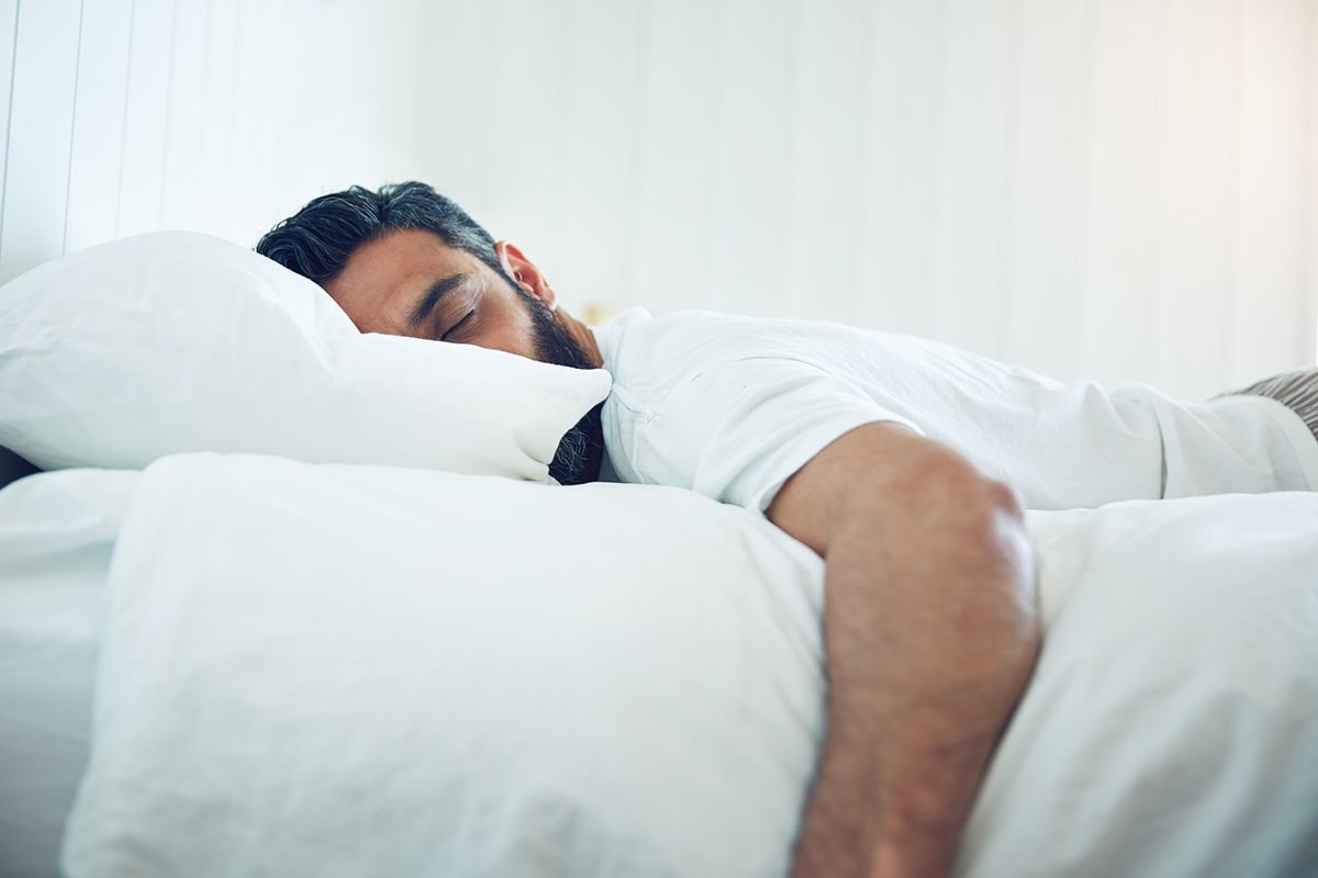 Brunette man with beard sound asleep face down on a bed with his arm hanging over the edge of the mattress. The pillowcase and sheets are white, as is the wall in the background.