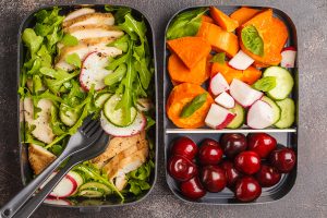 Two lunch boxes, one filled with grilled chicken on a bed of salad, the other with two compartments containing assorted veggies and cherries