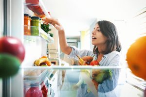 A woman looking through jars on the shelf of an open fridge door. Various produce in her arms and on the shelves.