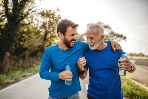Senior father and adult son, both in blue shirts, walking on an outdoor path. The younger man is holding a water bottle and has his arm around the older man. Both are laughing.