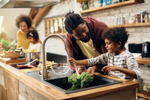 African American family prepping a meal in the kitchen. Father and son are in the foreground at the sink washing leafy greens, mother and daughter in the background prepping ingredients.