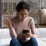 Confused angry woman looking at smartphone screen, sitting on couch at home, dissatisfied shocked young female having problem with discharged or broken device, reading spam message or bad news