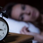 Close-up of alarm clock on night table, with woman lying awake on a white pillow blurred out in the background