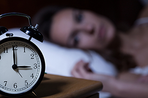 Close-up of alarm clock on night table, with woman lying awake on a white pillow blurred out in the background