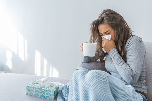Long-haired brunette woman sneezing into a kleenex while holding a white mug, with a blue blanket over her legs