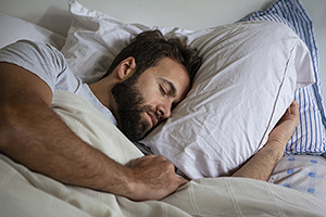Adult man with dark hair and beard asleep in bed with his arm under the pillow