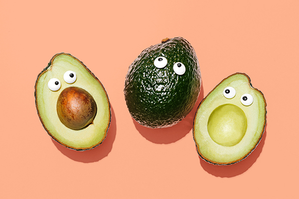 Two avocado halves and one whole avocado, each with a pair of googly eyes to make them look surprised, on a pastel peach background