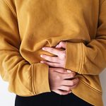 Woman in black leggings and ochre sweatshirt clutching her belly in discomfort