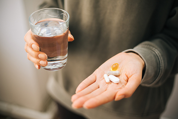 A caucasian person in a neutral colored sweatshirt holding vitamins and supplements being in an open palm, while their other hand holds a glass of water