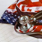 A stethoscope laying on top of a rumpled American flag over top of a graphic print paper of a cardiogram measuring heart beats
