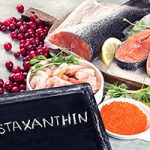Chalkboard with "astaxanthin" written on it, in front of astaxanthin-rich foods including salmon, salmon steaks, shrimp, fish eggs, and cranberries.