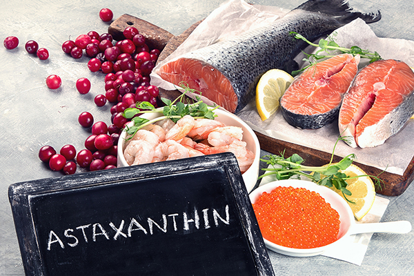 Chalkboard with "astaxanthin" written on it, in front of astaxanthin-rich foods including salmon, salmon steaks, shrimp, fish eggs, and cranberries.