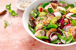 Salmon salad bowl with fresh radish, cucumber, red onion and green mixed leafy vegetables, in a white bowl on a stone countertop.