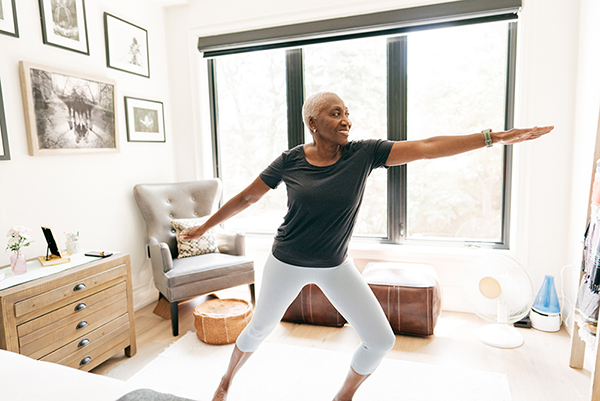 Senior African American woman doing yoga at home, in front of a large window.