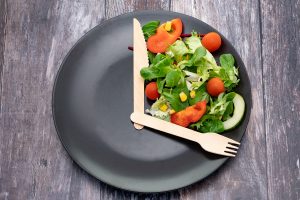 Black plate on a dark wooden background, with a fork and spoon positioned as the hands of a clock, a salad filling the space between 12 and 5.