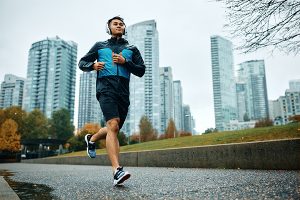 Man wearing headphones, black shorts, and a black and green jacket while running in a city park with tall buildings in the background