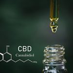 A dark background with the CBD chemical formula drawn in white next to the neck of an oil bottle beneath a dropper releasing a drop of CBD oil .