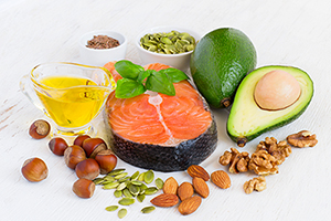 Array of foods with healthy fats, including olive oil, a salmon filet, an avocado, and a variety of nuts including almonds, hazelnuts, walnuts.