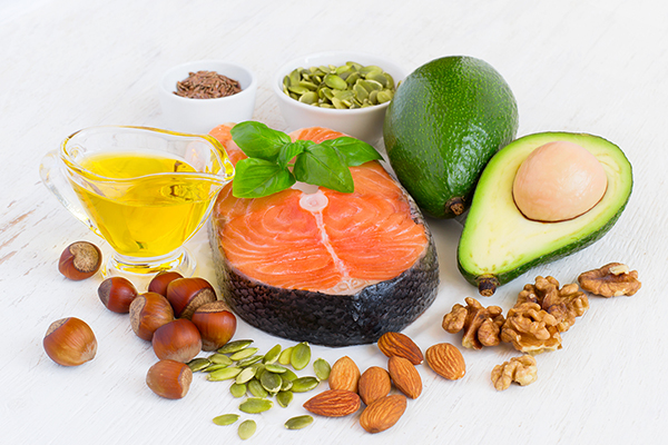 Array of foods with healthy fats, including a salmon filet, an avocado, and a variety of nuts including almonds, hazelnuts, walnuts.