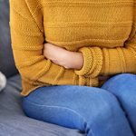 Closeup shot of a woman suffering with period cramps at home. She's sitting on a grey sofa and wearing blue jeans and a mustard-yellow sweater.