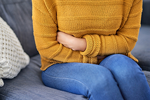 Closeup shot of a woman suffering with period cramps at home. She's sitting on a grey sofa and wearing blue jeans and a mustard-yellow sweater.