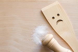 Wooden salt shaker lying on its side with salt spilled all around, next to a wooden spoon with a frowny face carved into it