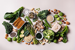 A variety of plant-based foods on a white background, including Tofu, soy beans, tempeh, green vegetables, nuts, seeds, quinoa, oats and spirulina.