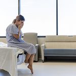 Young adult woman in hospital gown sitting on a hospital bed. She holds her face and cries after receiving some devastating health news.
