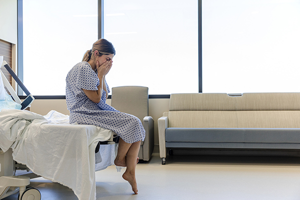 Young adult woman in hospital gown sitting on a hospital bed. She holds her face and cries after receiving some devastating health news.