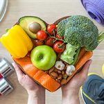 Two hands holding a heart-shaped dish full of healthy foods, including salmon, avocado, nuts, garlic, broccoli, garlic apples, and tomatoes. In the corners of the image, there are gym shoes, dumbells, and a yoga mat.