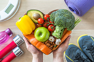 Two hands holding a heart-shaped dish full of healthy foods, including salmon, avocado, nuts, garlic, broccoli, garlic apples, and tomatoes. In the corners of the image, there are gym shoes, dumbells, and a yoga mat.