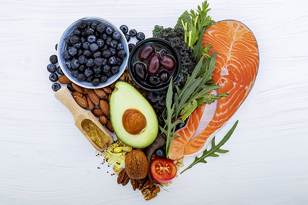 Heart shape of low carb foods on a white wooden background. Foods include blueberries, olives, salmon, avocado, greens, tomatoes, almonds, and some fish oil pills.