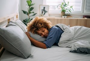 African American woman sleeping soundly on her stomach in the early morning. In the background, sun is coming though the window and shining on her houseplants.
