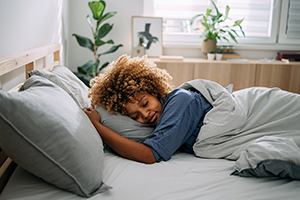 African American woman sleeping soundly on her stomach in the early morning. In the background, sun is coming though the window and shining on her houseplants.