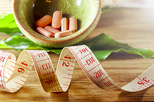 Weight measure tape coiled in foreground on a wooden table. In the background, a wooden bowl holds an assortment of supplements.