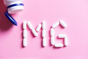 Pink background with an open pill bottle in the top left. Below it, white pill capsules spell out MG for magnesium.