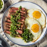 A white plate with blue trim resting on a folded cream-colored cloth napkin. The plate is half full of sliced skirt steak with chimichurri, the other half with two over-easy eggs.