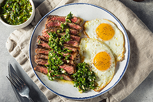 A white plate with blue trim resting on a folded cream-colored cloth napkin. The plate is half full of sliced skirt steak with chimichurri, the other half with two over-easy eggs.