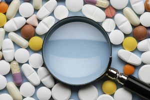Magnifying glass on blue background surrounded with an abundance of multicolored pills.
