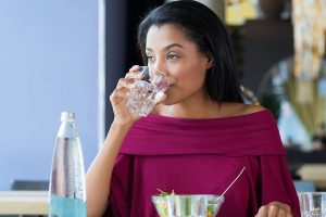 African American woman in purple off-the-shoulder top with a wide collar, drinking from a glass of water with a bowl of salad in front of her.